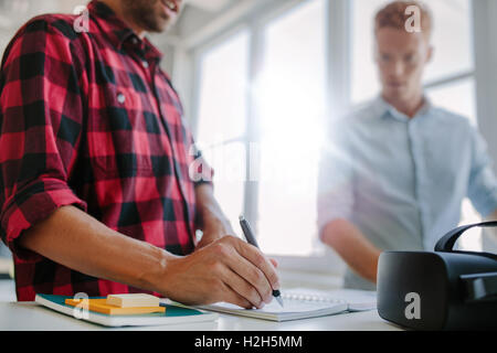 Closeup shot of young man writing on notepad, with colleague standing in background. Two young developers working together in of Stock Photo