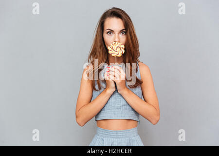 Funny cute young woman covered her mouth with lollipop over grey background Stock Photo