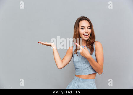 Smiling playful young woman winking and holding copyspace on palm over grey background Stock Photo