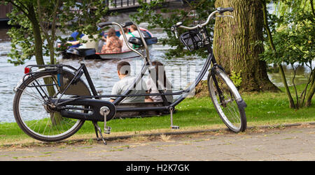 The tandem bicycle and back view of young couple relaxing near the water. Stock Photo