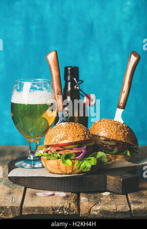 Two homemade beef burgers with crispy bacon, onion, pickles, vegetables, glass and bottle of wheat beer on wooden board over rus Stock Photo
