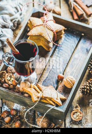 Glass of mulled wine in wooden tray with Christmas gingerbread cookies, nuts, cinnamon, anise and pine cones over rustic wooden Stock Photo