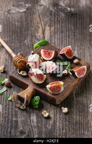 Fresh sliced figs with ricotta cheese, basil leaves, hazelnut and honey from honey dipper on small wood cutting board over dark Stock Photo