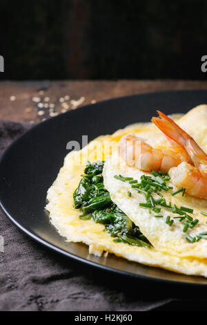 Omelet with cooked spinach and fried shrimps prawns, served with cutting chive and sesame seeds on black plate with textile napk Stock Photo