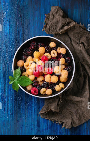 Bowl of colorful yellow and red raspberries and black dewberry with leaf on sackcloth rag over blue wooden textured background. Stock Photo