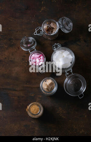 Different glass jars with variety of sugar white, brown pink fruit granulated, cubes and crystal over old dark wood background. Stock Photo