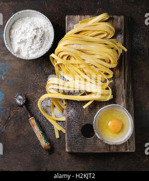 Raw uncooked homemade italian pasta tagliatelle with, pasta cutter, bowls with white flour and broken egg on old wood cutting bo Stock Photo