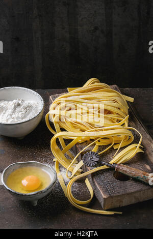 Raw uncooked homemade italian pasta tagliatelle with, pasta cutter, bowls with white flour and broken egg on old wood cutting bo Stock Photo