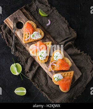 Wafers as heart shape with salted salmon, red onion, chive, lime and ricotta cheese on wooden cutting board with sackcloth rag o Stock Photo