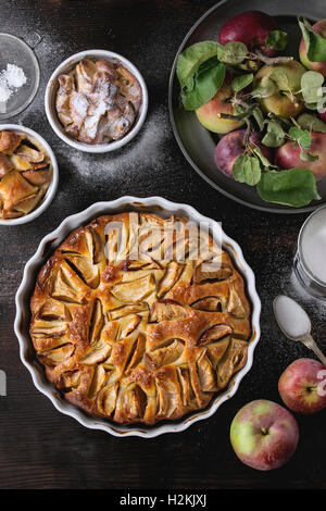 Homemade big and small apple cake pie in white ceramic forms with fresh apples with leaves, sugar powder, and tin can of sugar o Stock Photo