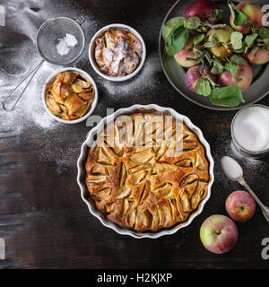 Homemade big and small apple cake pie in white ceramic forms with fresh apples with leaves, sugar powder, and tin can of sugar o Stock Photo