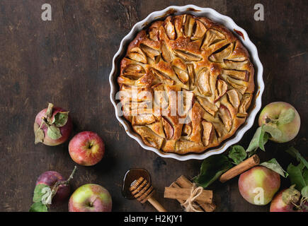 Homemade apple cake pie in white ceramic form with fresh apples with leaves, honey and cinnamon sticks over dark wooden backgrou Stock Photo