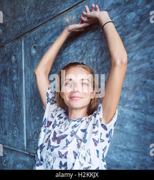 Pretty blond woman standing against grey brick wall with raised hands smiling and looking at the camera Stock Photo