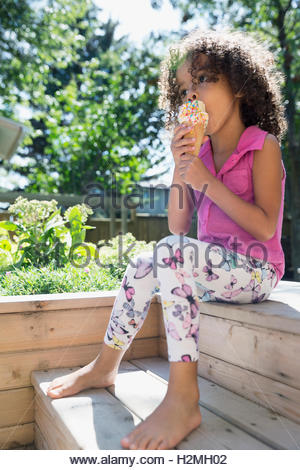 Barefoot girl eating ice cream cone with sprinkles in sunny backyard