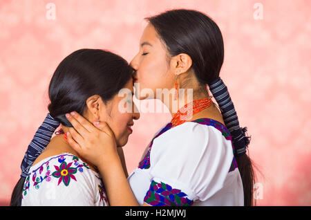 Beautiful hispanic mother and daughter wearing traditional andean clothing, seen from profile angle facing each other, young woman kissing her mom on forehead, pink studio background Stock Photo