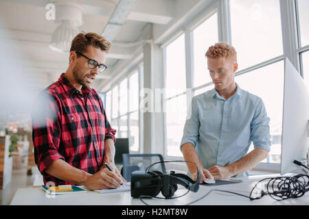 Two young businessman working together. They are standing at a table with virtual reality glasses. One using computer and other Stock Photo