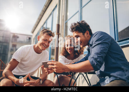 Three young friends sitting outdoors and looking at mobile phone. Group of people sitting at outdoor cafe and watching video on Stock Photo