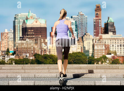 Rear view of a slim woman running up stairs on a sunny day. The background shows the blurred skyline of Brooklyn New York. Stock Photo