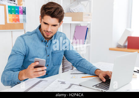 Smiling young businessman in glasses using laptop and smartphone at workplce Stock Photo