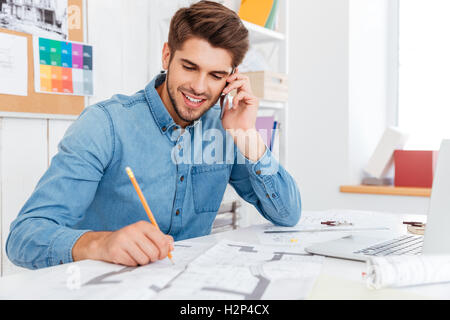 Smiling young businessman talking on mobile phone and making notes while sitting at the office Stock Photo