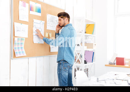 Young casual businessman looking at office task board and talking on the phone Stock Photo
