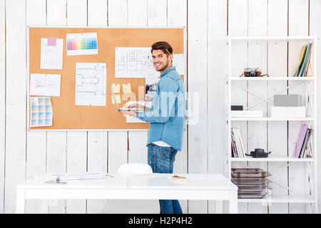 Smiling young man holding laptop and standing at task board in office Stock Photo