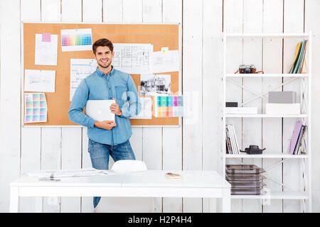 Cheerful young man standing at the task board and holding laptop in office Stock Photo