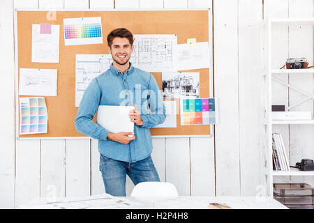 Cheerful happy young man standing at the task board and holding laptop in office Stock Photo