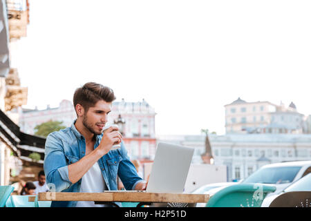 Smiling cheerful casual man using laptop while sitting at cafe outdoors and drinking coffee Stock Photo