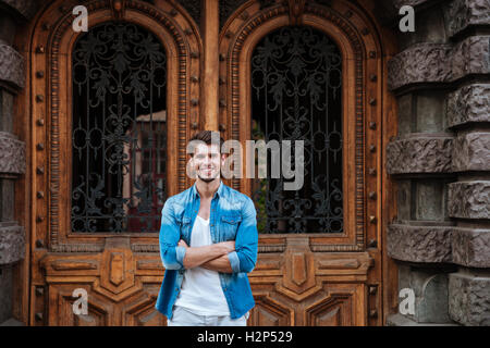 Handsome young smiling man standing in front of the beautiful wooden door waiting for someone outdoors Stock Photo