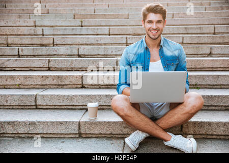 Young casual smiling man student using laptop while sitting on the staircase outdoors with cup of coffee Stock Photo