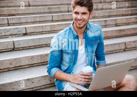 Cheerful young man with laptop drinking take away coffee while sitting on the staircase outdoors Stock Photo
