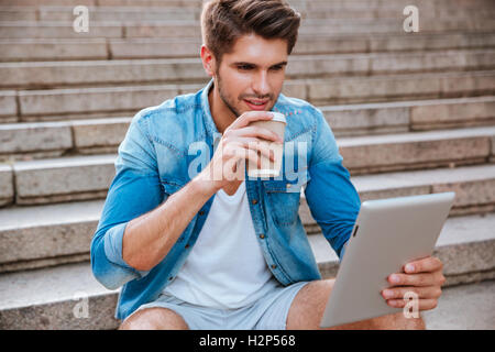 Confident young man drinking take away coffee and looking at pc tablet while sitting on the staircase outdoors