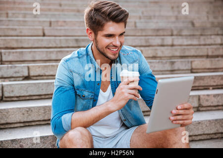 Smiling happy casual man using pc tablet and drinking coffee outdoors on the staircase Stock Photo