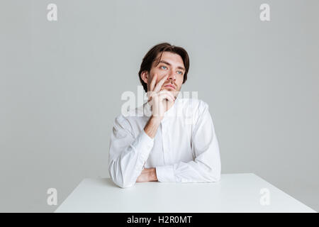 Thoughtful young man thinking about something while sitting at the table isolated on the gray background Stock Photo