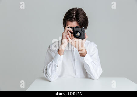 Handsome young brunette man sitting at the table and making photo with camera isolated on the gray background Stock Photo