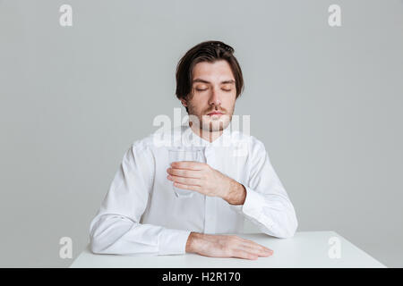 Relaxed young man holding water glass while sitting at the table isolated on the gray background Stock Photo