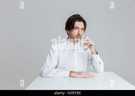 Handsome young man drinking from water glass while sitting at the table isolated on the gray background Stock Photo