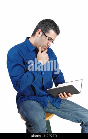 A young man in a blue white and jeans sitting whit a book in his hand and glasses, reading in his book, for white background. Stock Photo