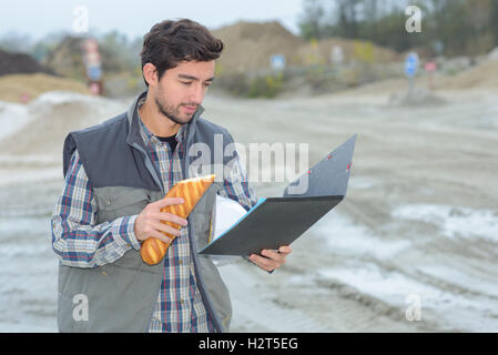 Man on building site looking at file and eating baguette Stock Photo