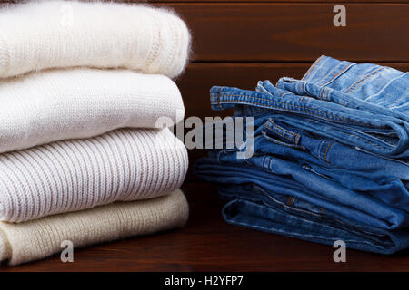 Stack of white and cozy knitted sweaters and denim jeans on rustic wooden background Stock Photo