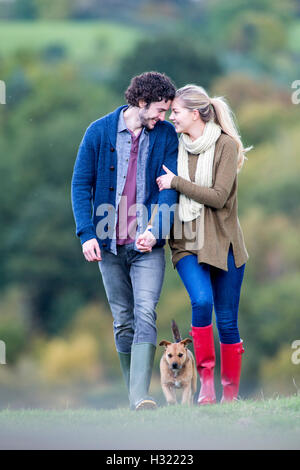 Young couple walking together on a rainy day with their pet dog. They have their heads together and are holding hands. Stock Photo