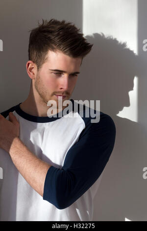 Portrait of an attractive young man, standing against a white wall with sunlight from the window shining on him. Stock Photo