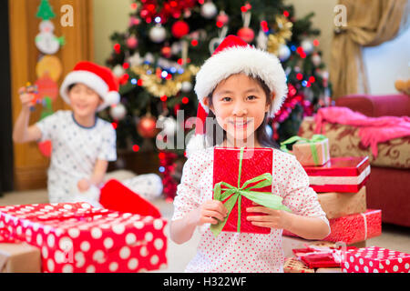 Happy Chinese girl sitting on the floor in her living room at Christmas time. She is surrounded by presents, one of which she is Stock Photo