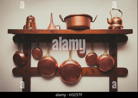 Different kind of vintage copper cookware, pans, coffee pot and funnel hanging on wooden shelf in kitchen with white rough wall Stock Photo