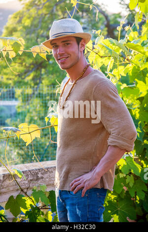 Side view of handsome young man in hat toching vine leaves in garden in sunlight Stock Photo