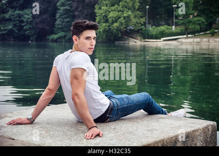 Contemplative light brown haired handsome young man wearing white t-shirt and denim jeans sitting on wall beside picturesque riv Stock Photo