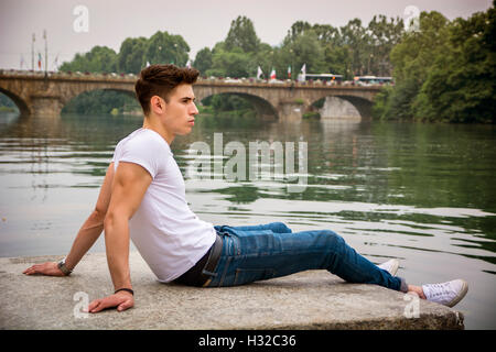 Contemplative light brown haired handsome young man wearing white t-shirt and denim jeans sitting on wall beside picturesque riv Stock Photo