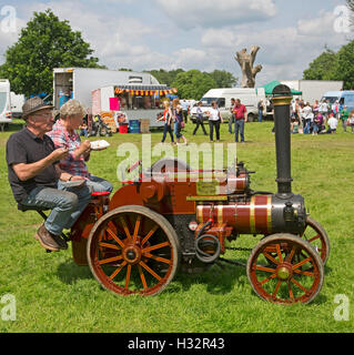 Man & woman on seat of miniature steam traction engine eating takeaway food at English country fair with food van in background Stock Photo