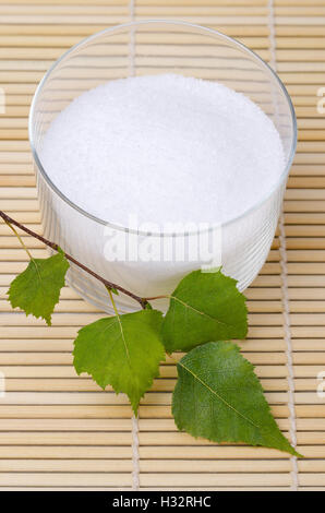 Xylitol birch sugar in a glass bowl with birch leaves on a bamboo mat. White granulated sugar alcohol, a sweetener. Stock Photo